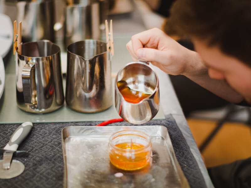 A hand pouring glowing orange melted wax from a stainless steel jug into a glass ramekin